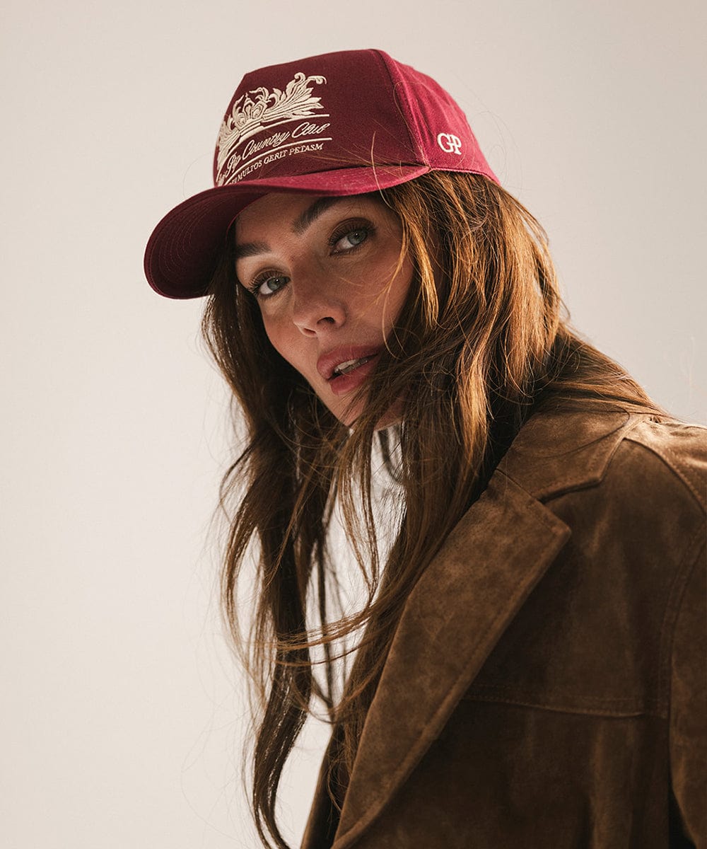 Woman wearing a burgundy vintage trucker cap with a white logo and a brown coat against a plain background #color_burgundy