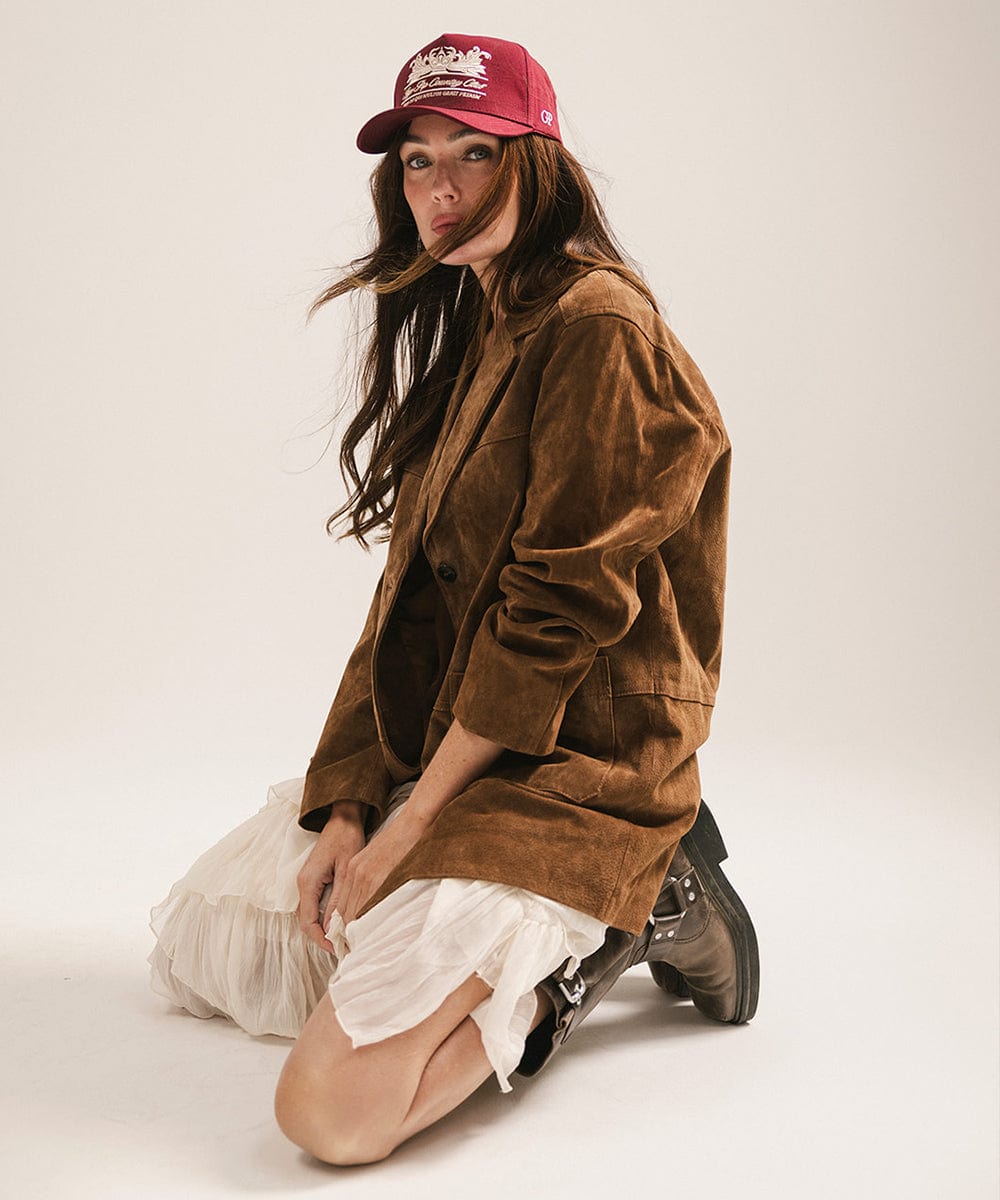 Woman wearing a burgundy vintage trucker cap with a white logo, a brown coat and a white skirt, sitting against a plain background #color_burgundy