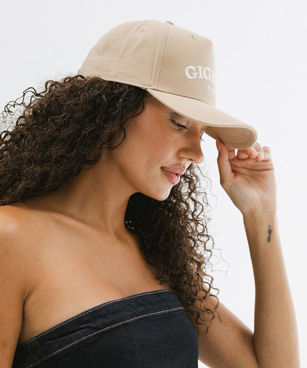 Woman wearing a tan trucker hat with 'GIGI PIP' embroidered in white on the front and a denim top, posing sideways against a plain background #color_tan