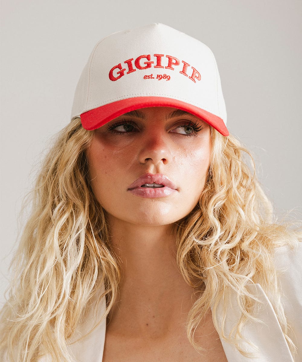 Woman wearing a cream and red trucker hat with 'GIGI PIP' embroidered in red on the front against a plain background #color_cream - red