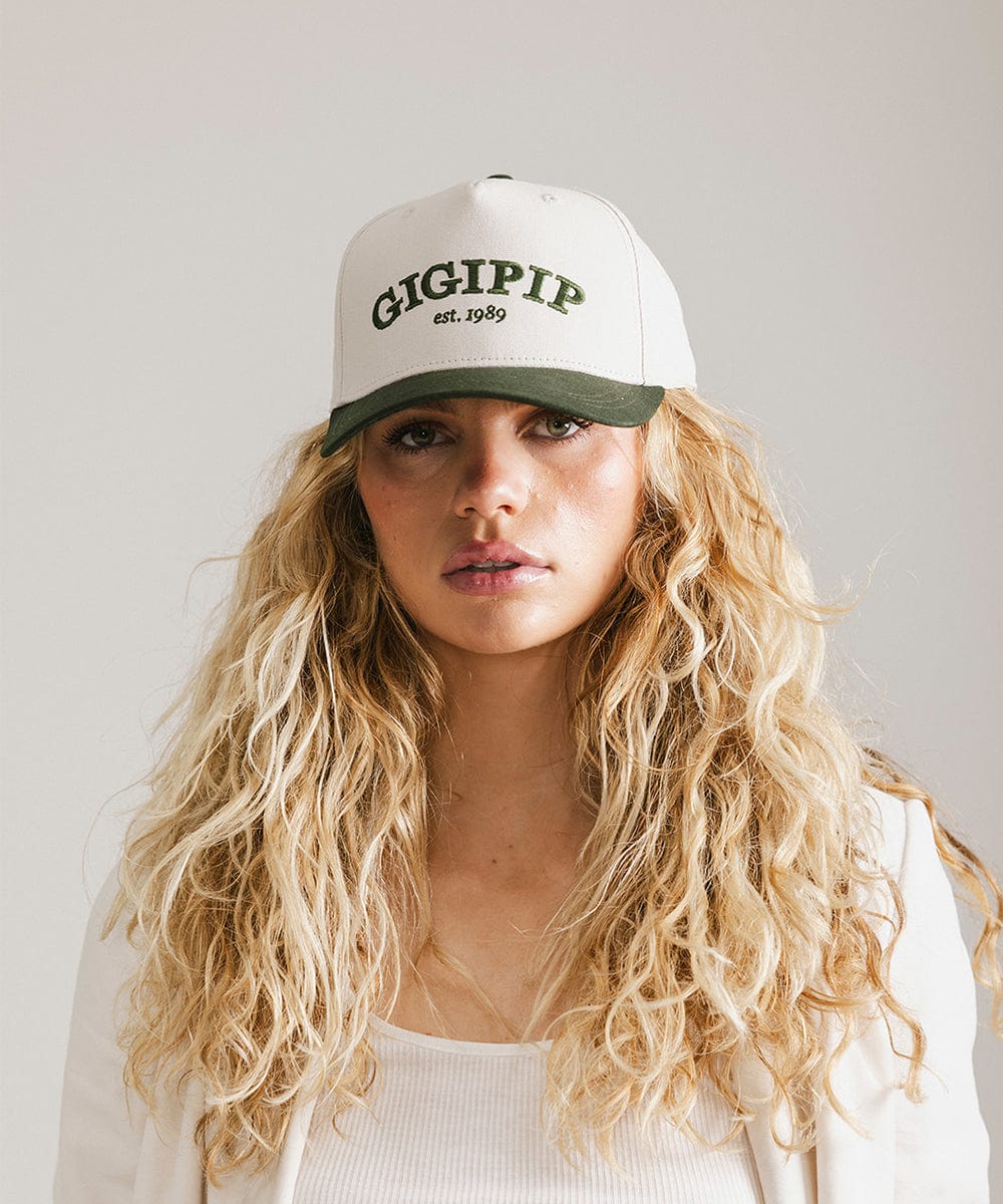 Woman wearing a cream and dark green trucker hat with 'GIGI PIP' embroidered in dark green on the front and a white coat against a plain background #color_cream - dark green