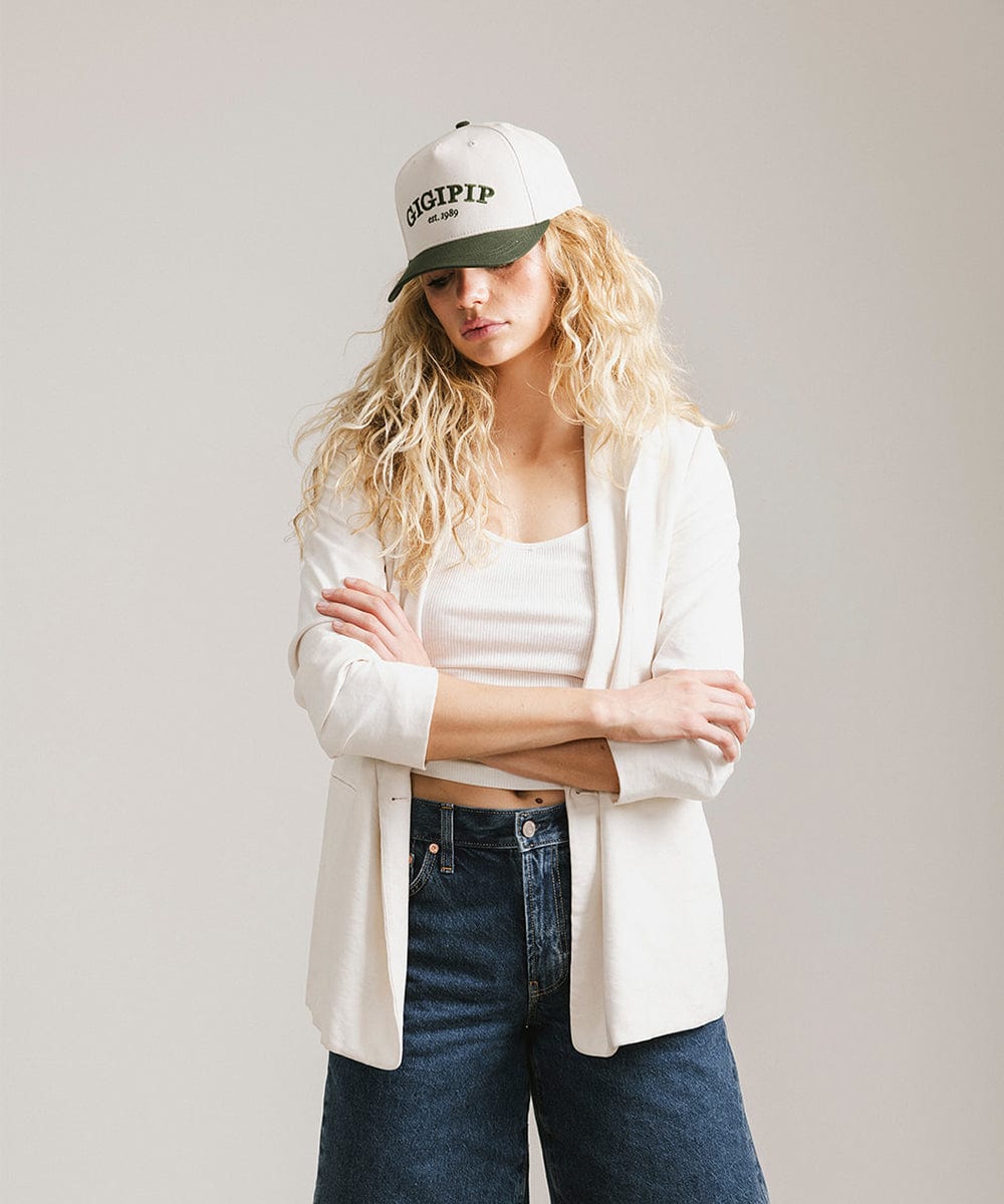 Woman wearing a cream and dark green trucker hat with 'GIGI PIP' embroidered in dark green on the front, a white top with a white coat and blue jeans, posing against a plain background #color_cream - dark green