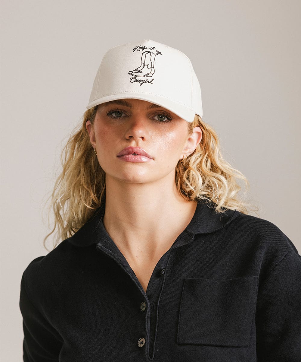 Woman wearing a cream cap with black text and boot design, and a black shirt against a plain background #color_cream