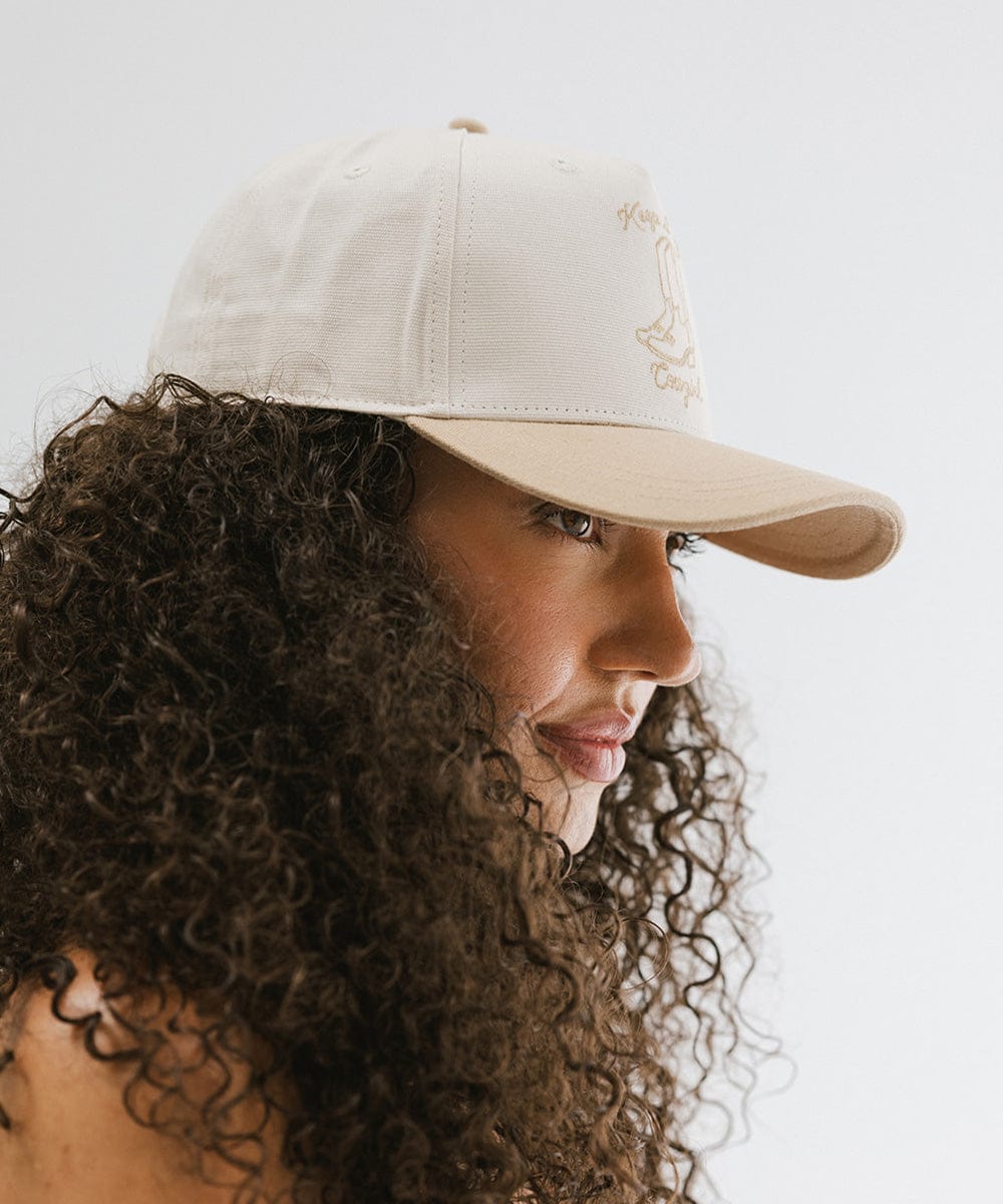 Woman with curly hair wearing a cream and tan cap with tan text and boot design, posing sideways against a plain background #color_cream - tan