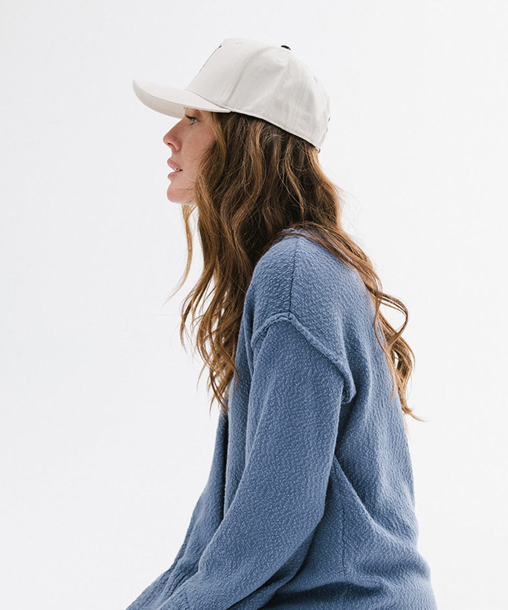 Woman wearing a cream cap with black text and boot design, and a blue sweater, facing sideways against a plain background #color_cream