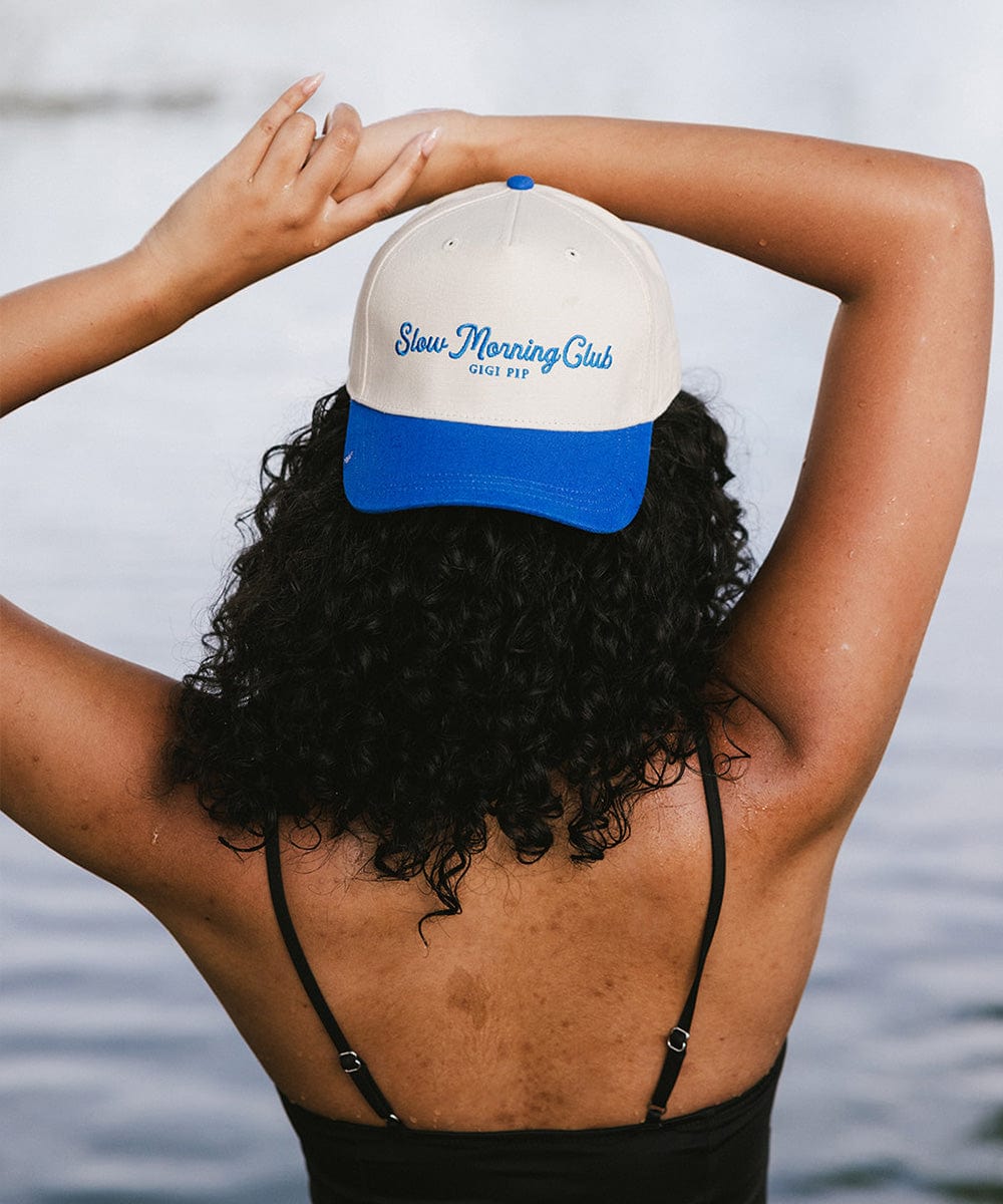 Person with curly hair wearing a cream and blue cap with 'Slow Morning Club' text and a black top, facing the sea in the background #color_cream - blue