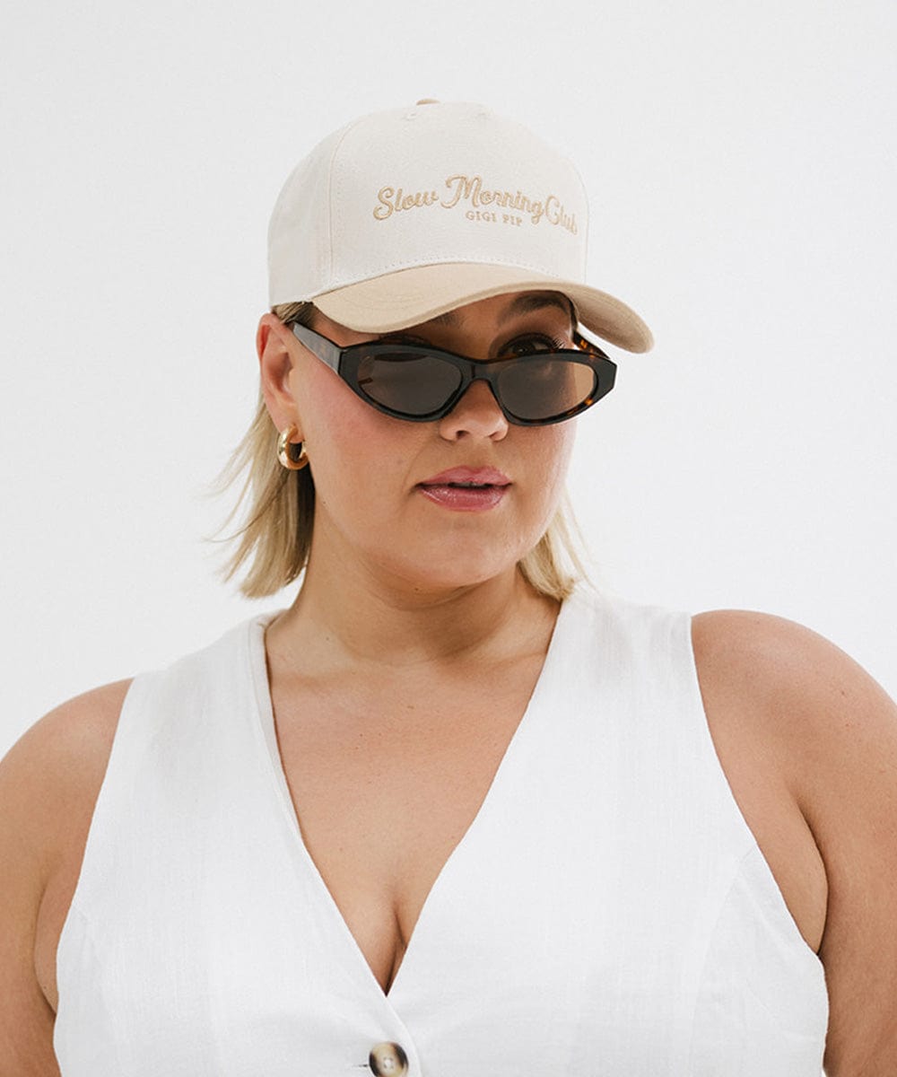 Woman wearing a cream and tan cap with 'Slow Morning Club' text, sunglasses, and a white top against a plain background #color_cream - tan