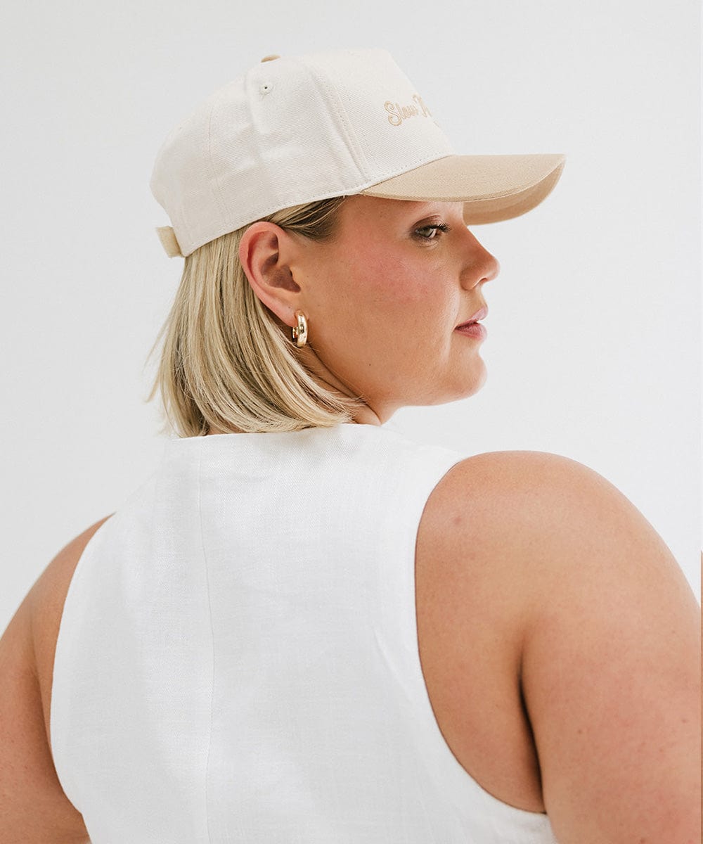 Woman wearing a cream and tan cap with 'Slow Morning Club' text and a white top, facing sideways against a plain background #color_cream - tan