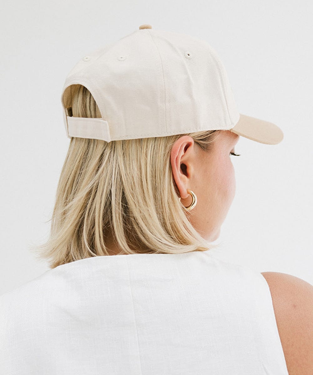 Woman wearing a cream and tan cap with 'Slow Morning Club' text, sunglasses, and a white top, facing behind against a plain background #color_cream - tan