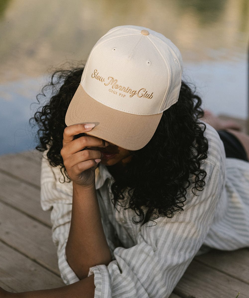 Woman wearing a cream and tan cap with 'Slow Morning Club' text and a light colored shirt, lying down against a natural background #color_cream - tan