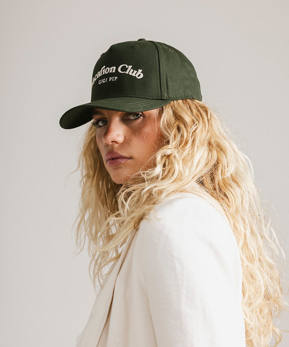 Woman wearing a dark green cap with 'Vacation Club' text and a white coat, posing sideways against a plain background #color_dark green