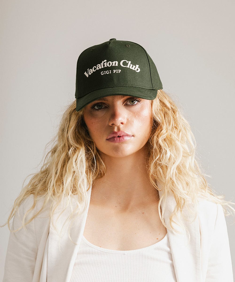 Woman wearing a dark green cap with 'Vacation Club' text and a white coat against a plain background #color_dark green