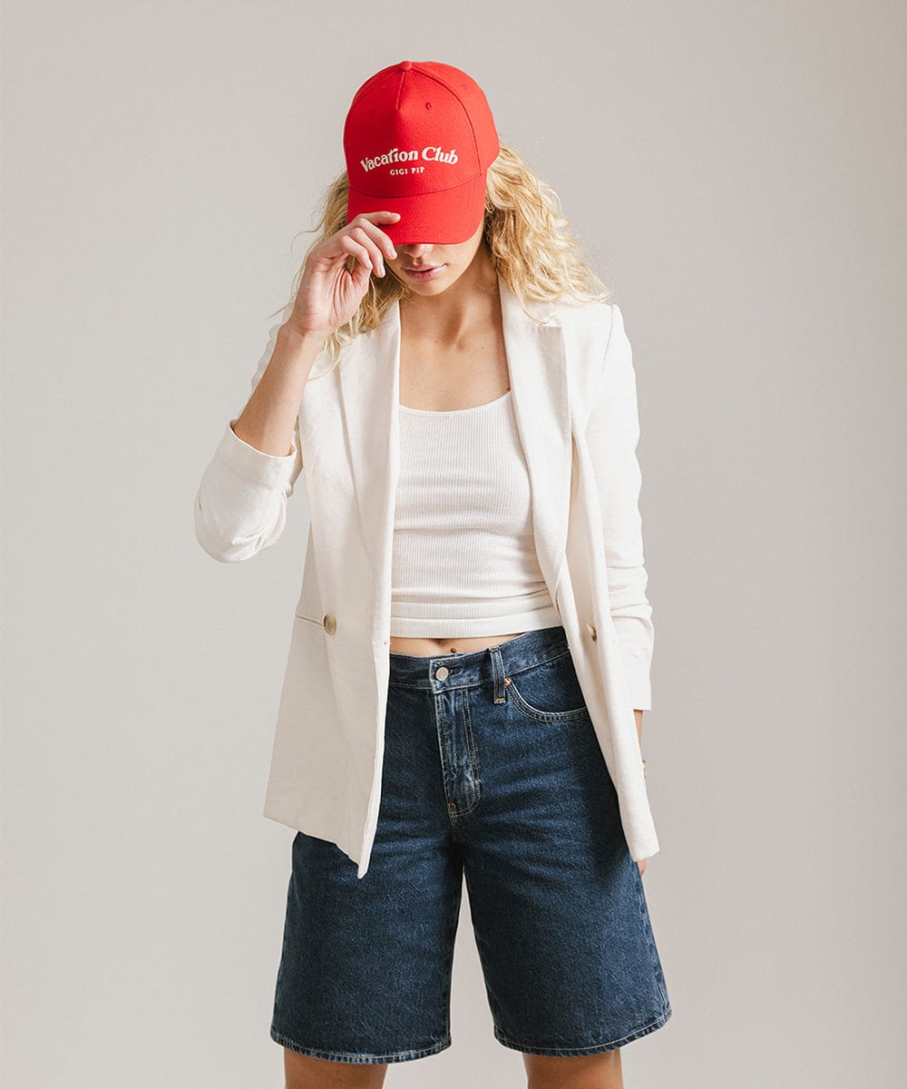 Woman wearing a red cap with 'Vacation Club' text, a white top with a white coat and denim shorts, posing against a plain background #color_red