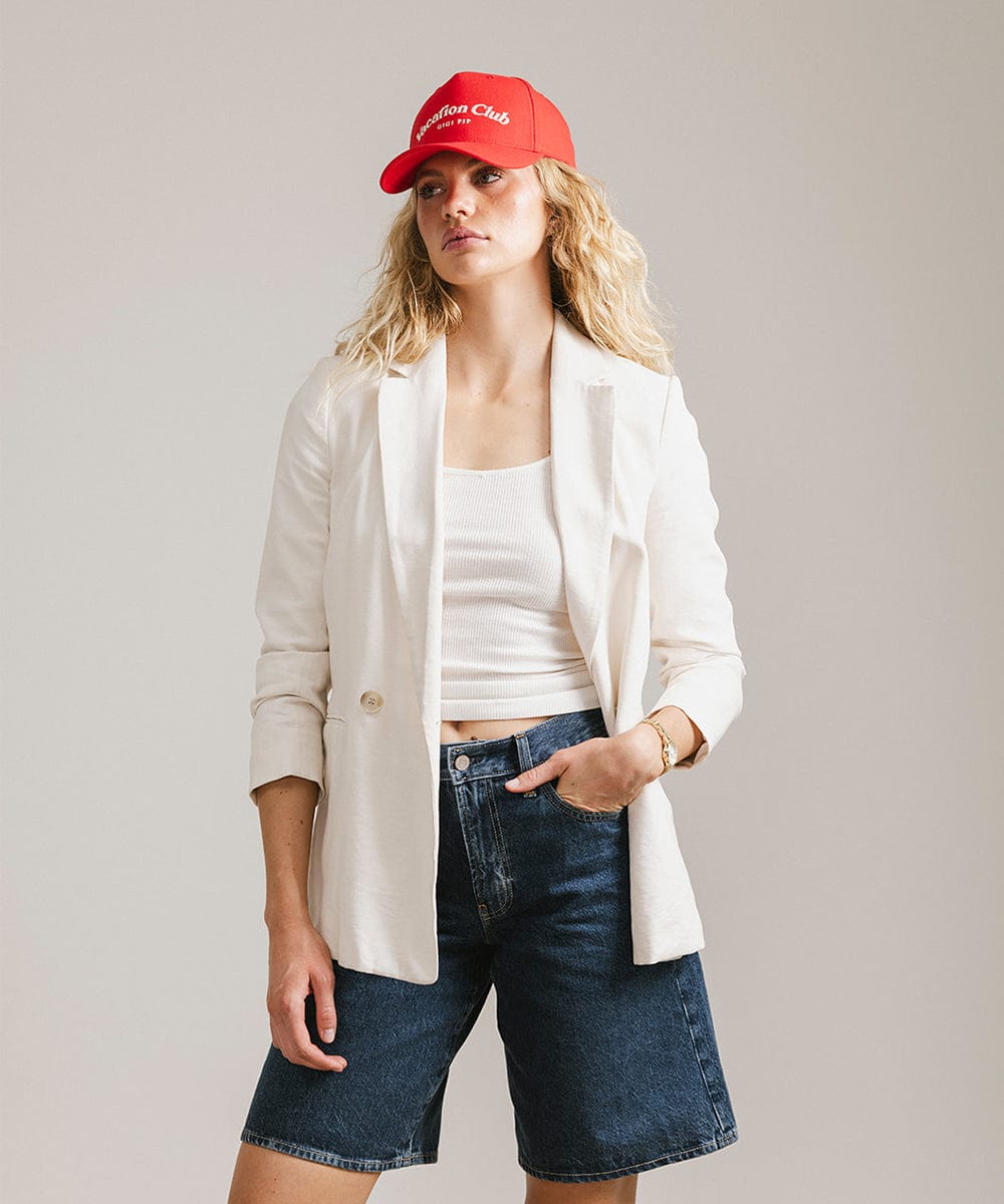 Woman wearing a red cap with 'Vacation Club' text, a white top with a white coat and denim shorts against a plain background #color_red
