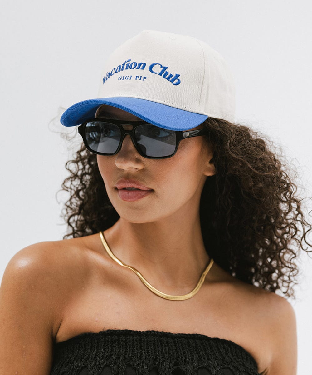 Woman wearing a cream and blue cap with 'Vacation Club' text, sunglasses and a gold chain, posing against a plain background #color_cream - blue