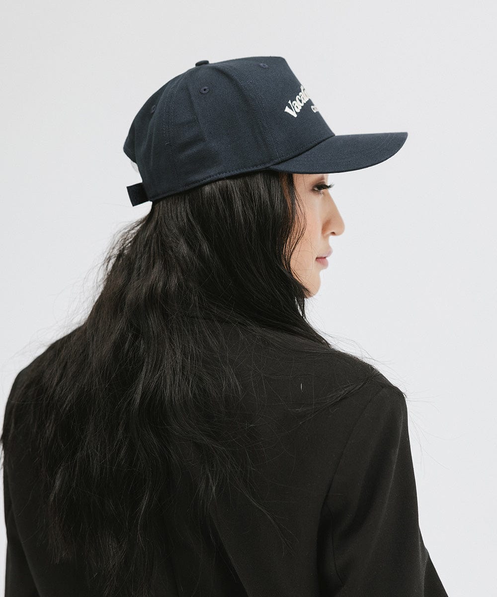 Woman wearing a navy cap and a black blazer, facing sideways against a plain background #color_navy