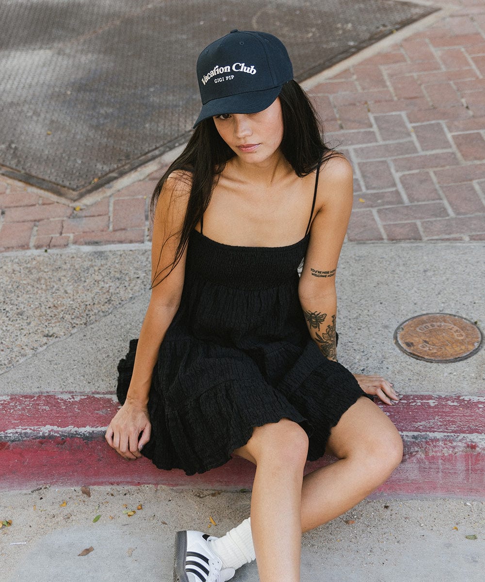 Woman wearing a navy cap with 'Vacation Club' text and a black dress, sitting on a pavement #color_navy