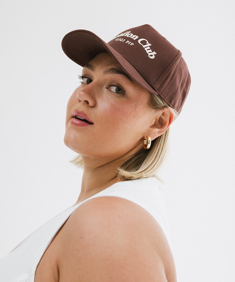 Woman wearing a chocolate brown colored cap with 'Vacation Club' text and a white top, posing sideways against a plain background #color_chocolate brown
