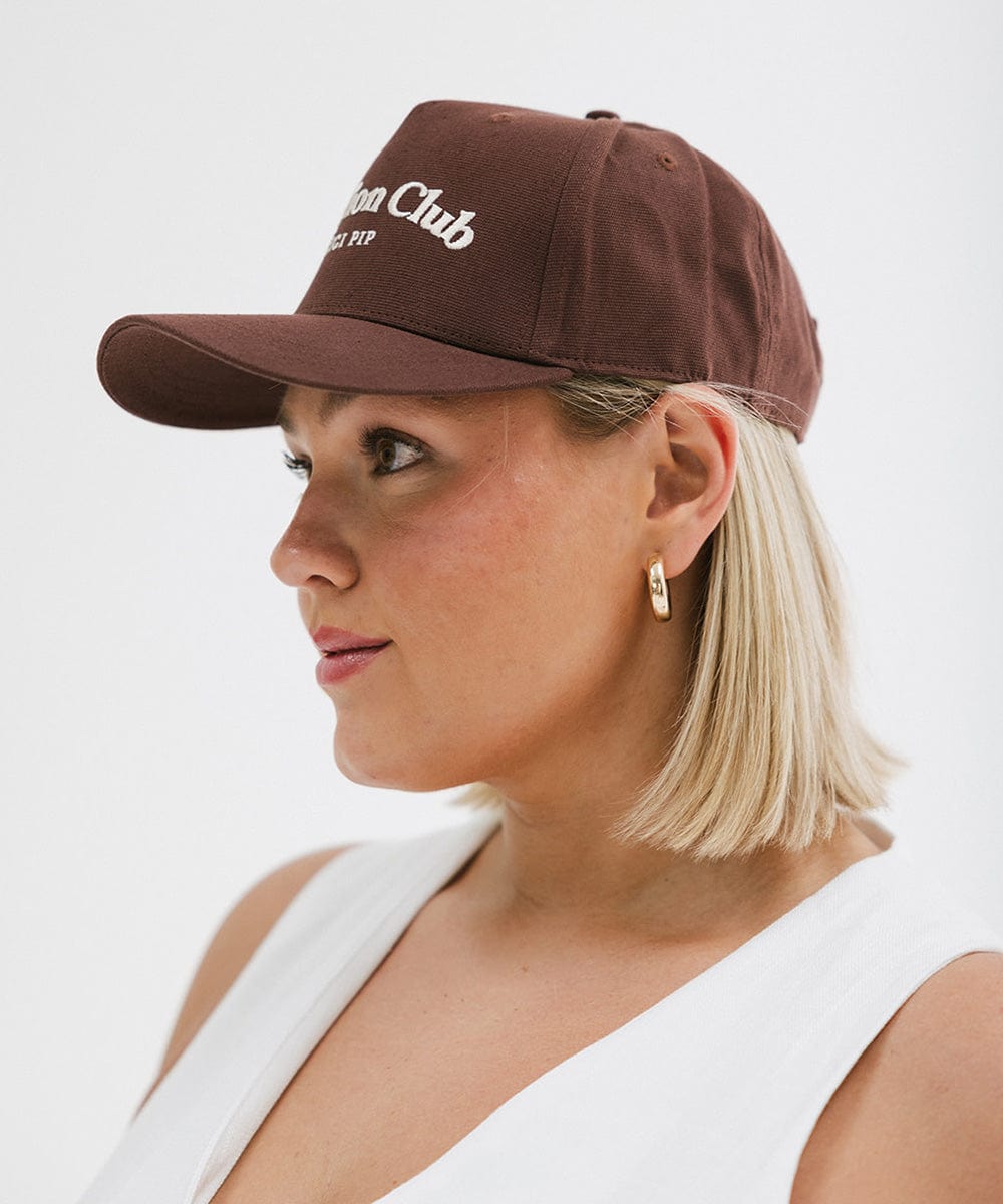 Woman wearing a chocolate brown colored cap with 'Vacation Club' text and a white top, facing sideways against a plain background #color_chocolate brown