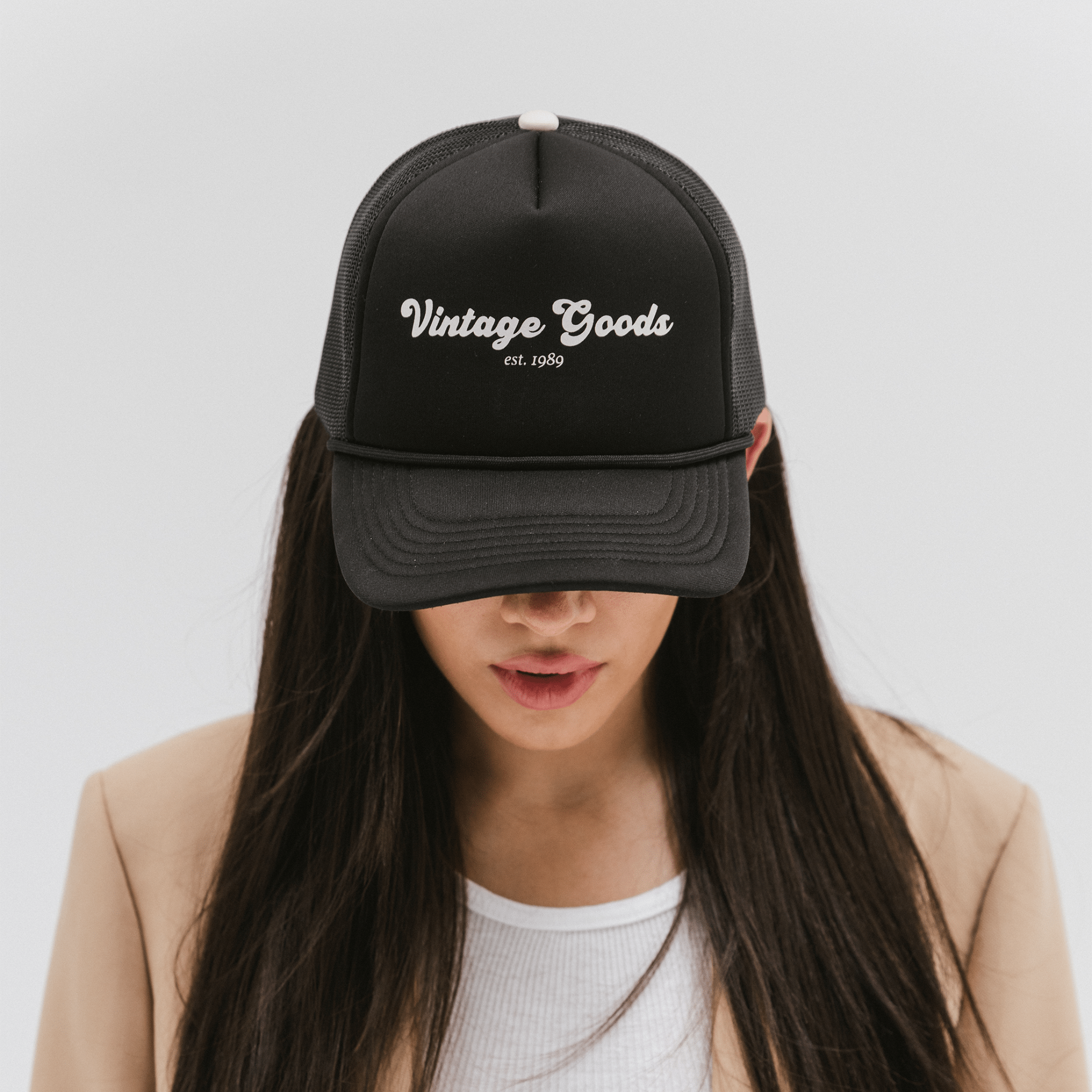 Woman wearing a black trucker hat with 'Vintage Goods' text and a beige blazer, looking down against a plain background #color_black