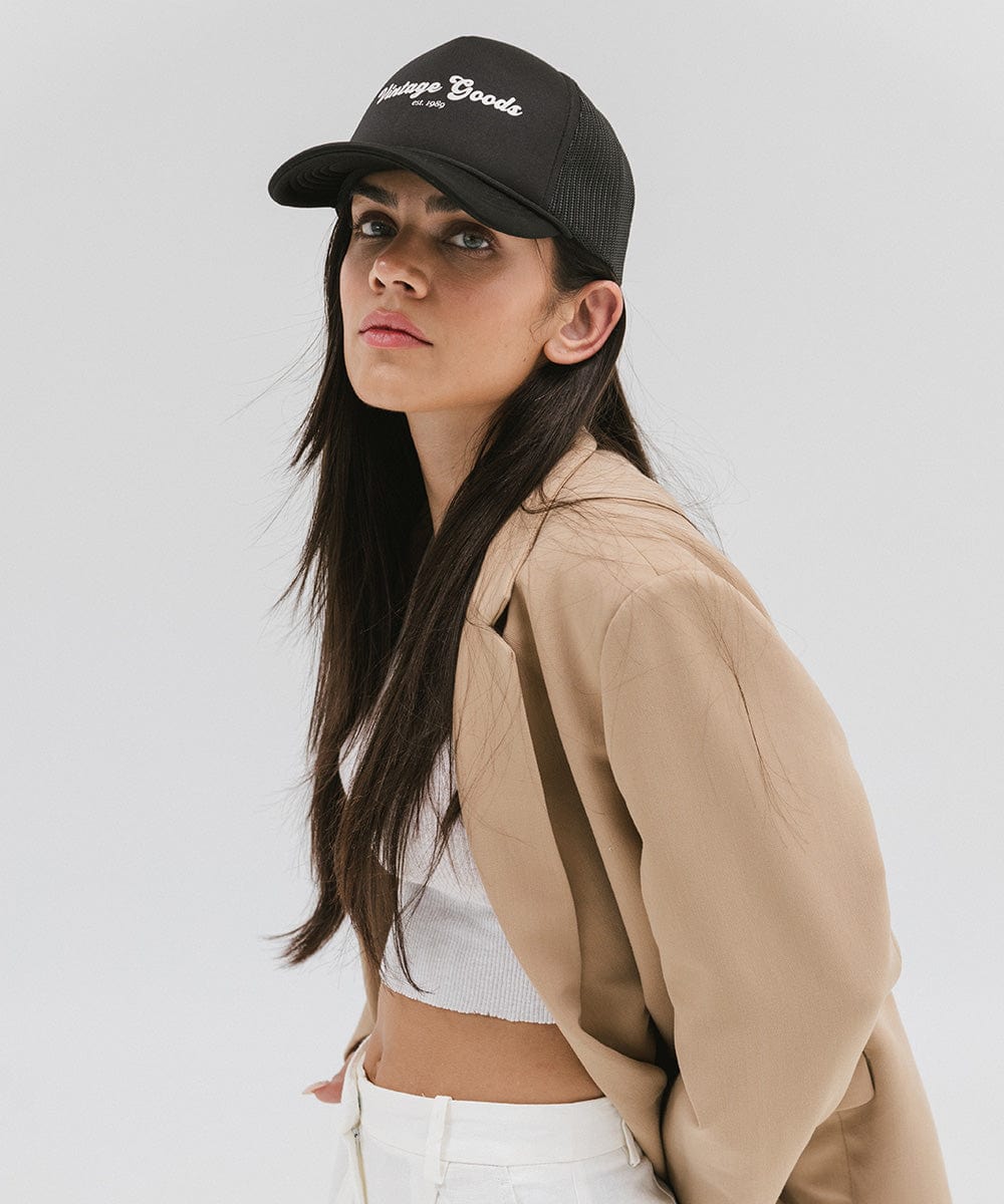 Woman wearing a black trucker hat with 'Vintage Goods' text and a beige blazer, posing against a plain background #color_black