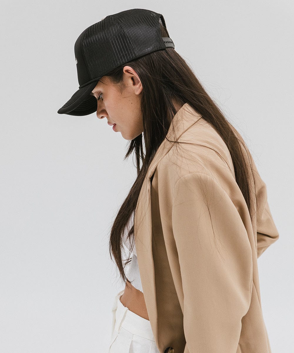 Woman wearing a black trucker hat with 'Vintage Goods' text and a beige blazer, facing sideways against a plain background #color_black