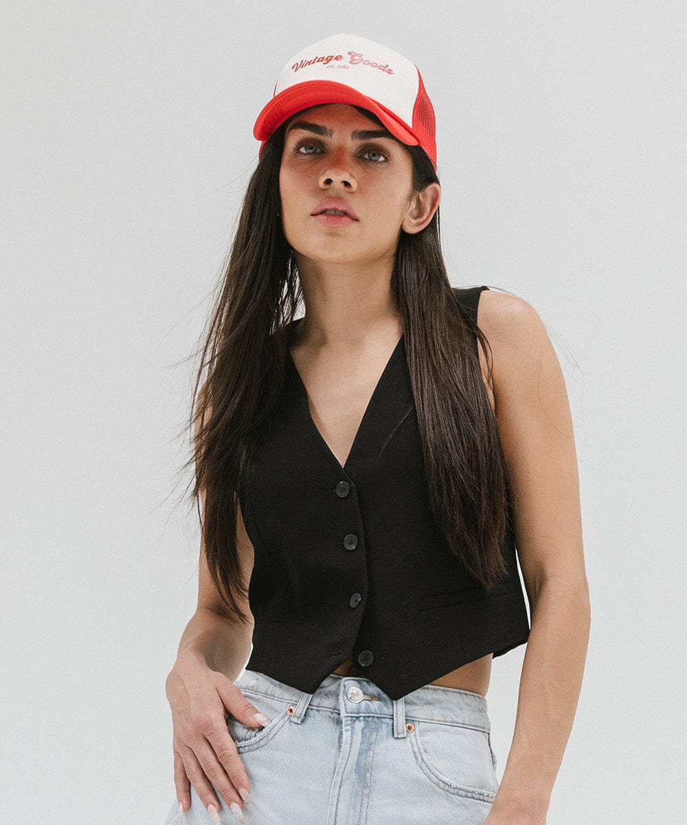 Woman wearing a cream and vintage red colored trucker hat with 'Vintage Goods' text, a black top and light blue jeans against a plain background #color_cream-vintage red