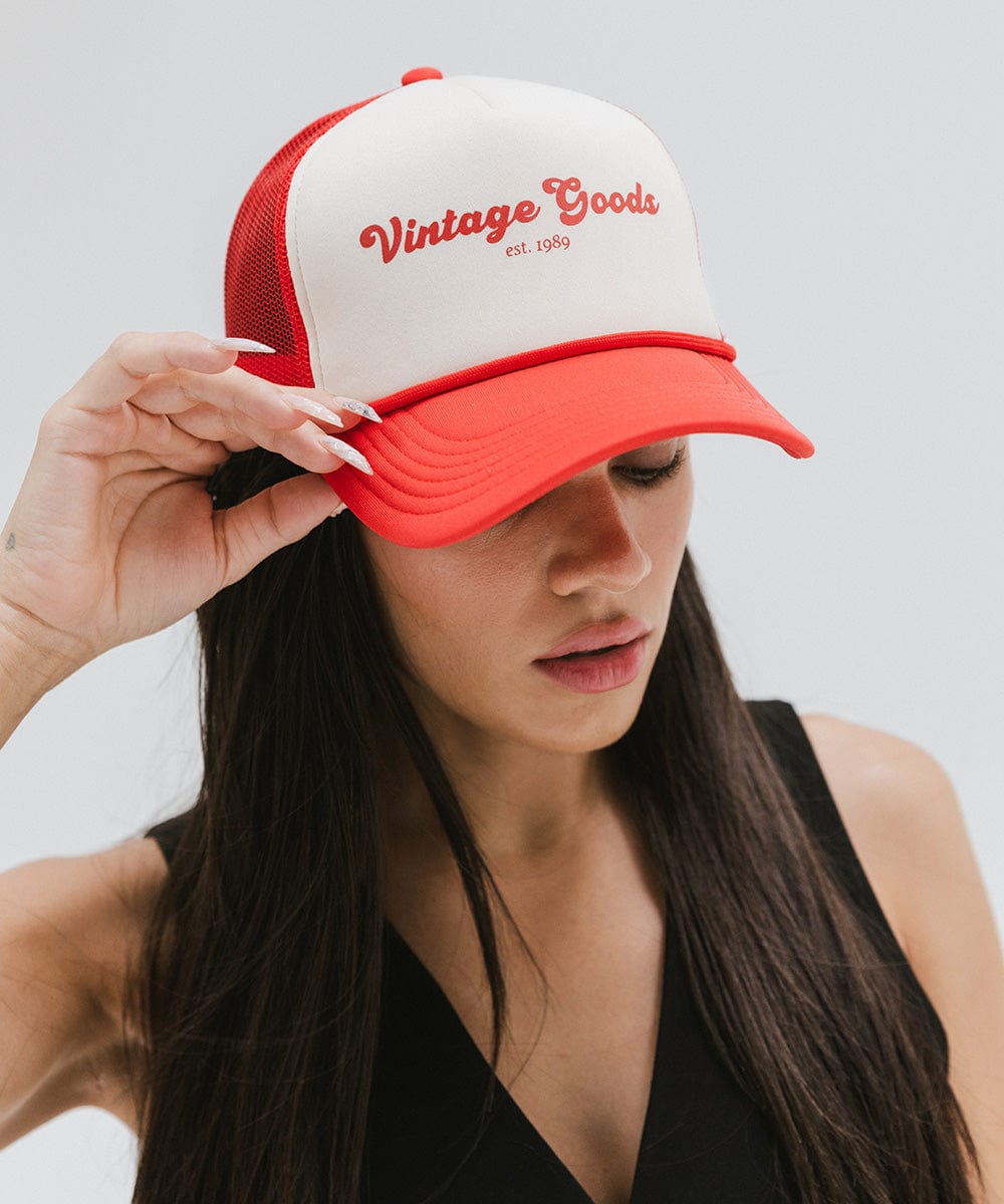 Woman wearing a cream and vintage red colored trucker hat with 'Vintage Goods' text and a black top, looking sideways against a plain background #color_cream-vintage red