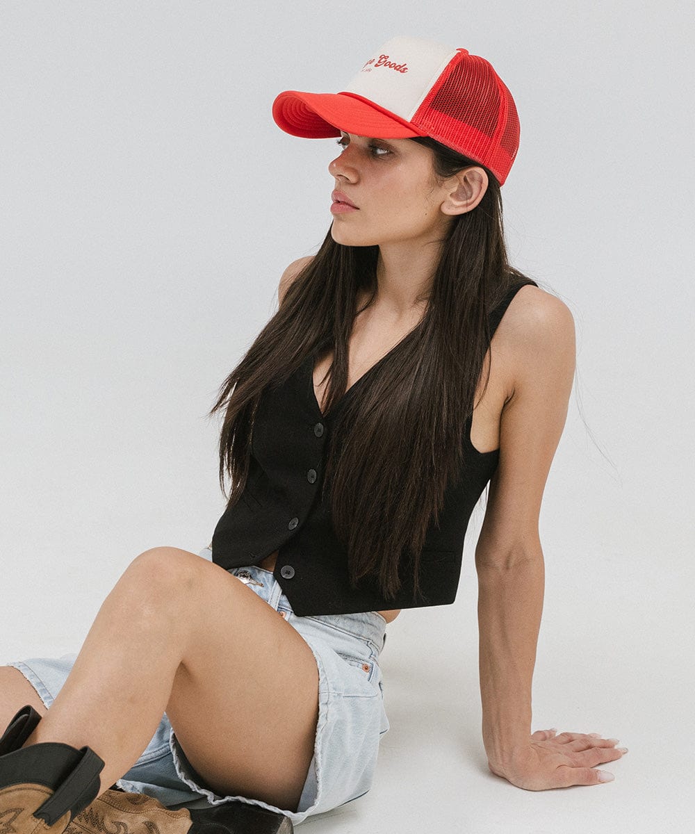Woman wearing a cream and vintage red colored trucker hat with 'Vintage Goods' text, a black top, denim shorts and brown boots, sitting against a plain background #color_cream-vintage red