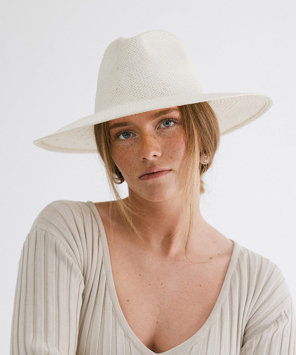 Woman wearing a white wide brim fedora straw hat and a grey top against a plain background  #color_white
