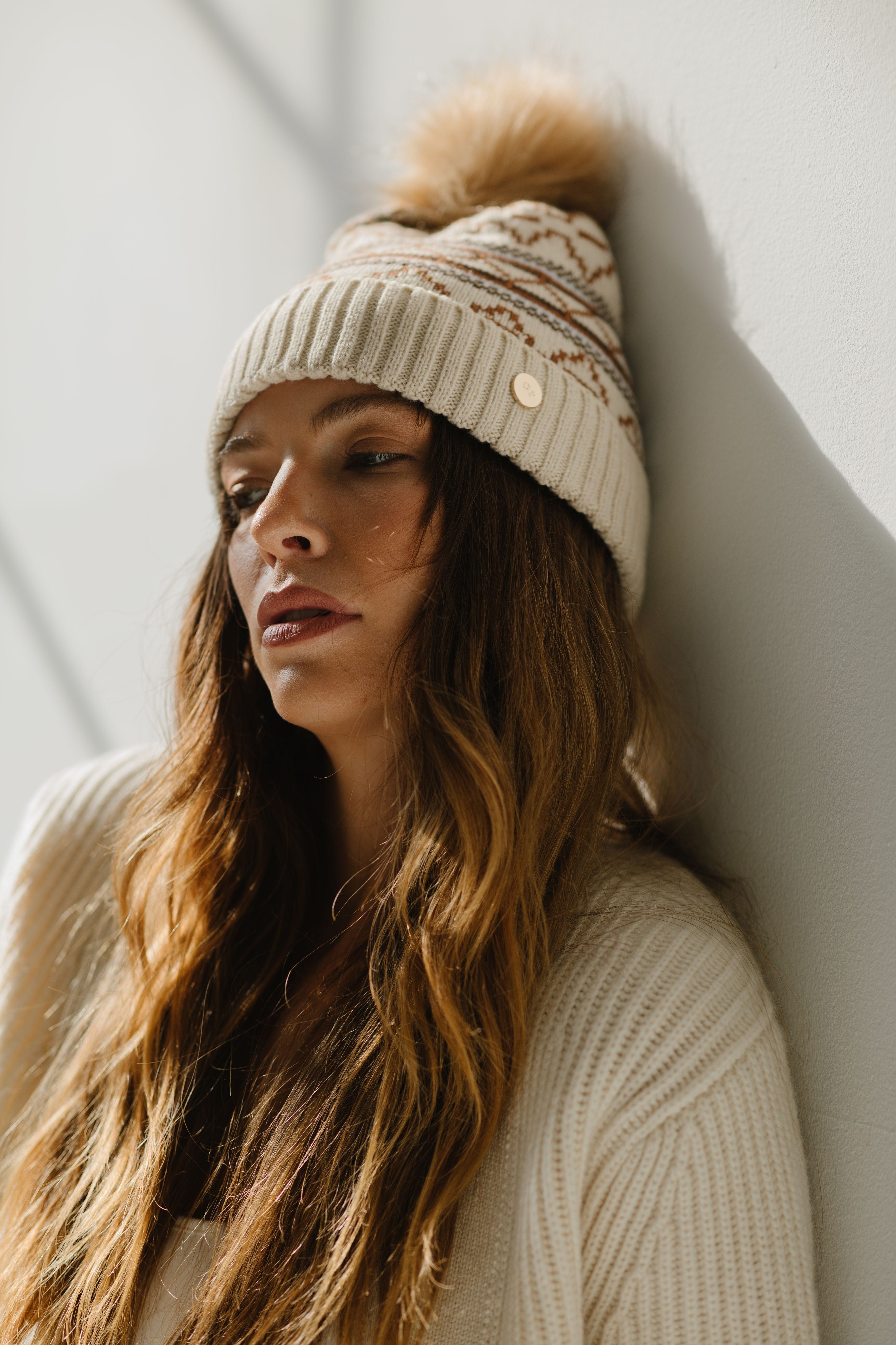 Woman wearing a printed knit beanie and a white sweater, posing against a wall in the background