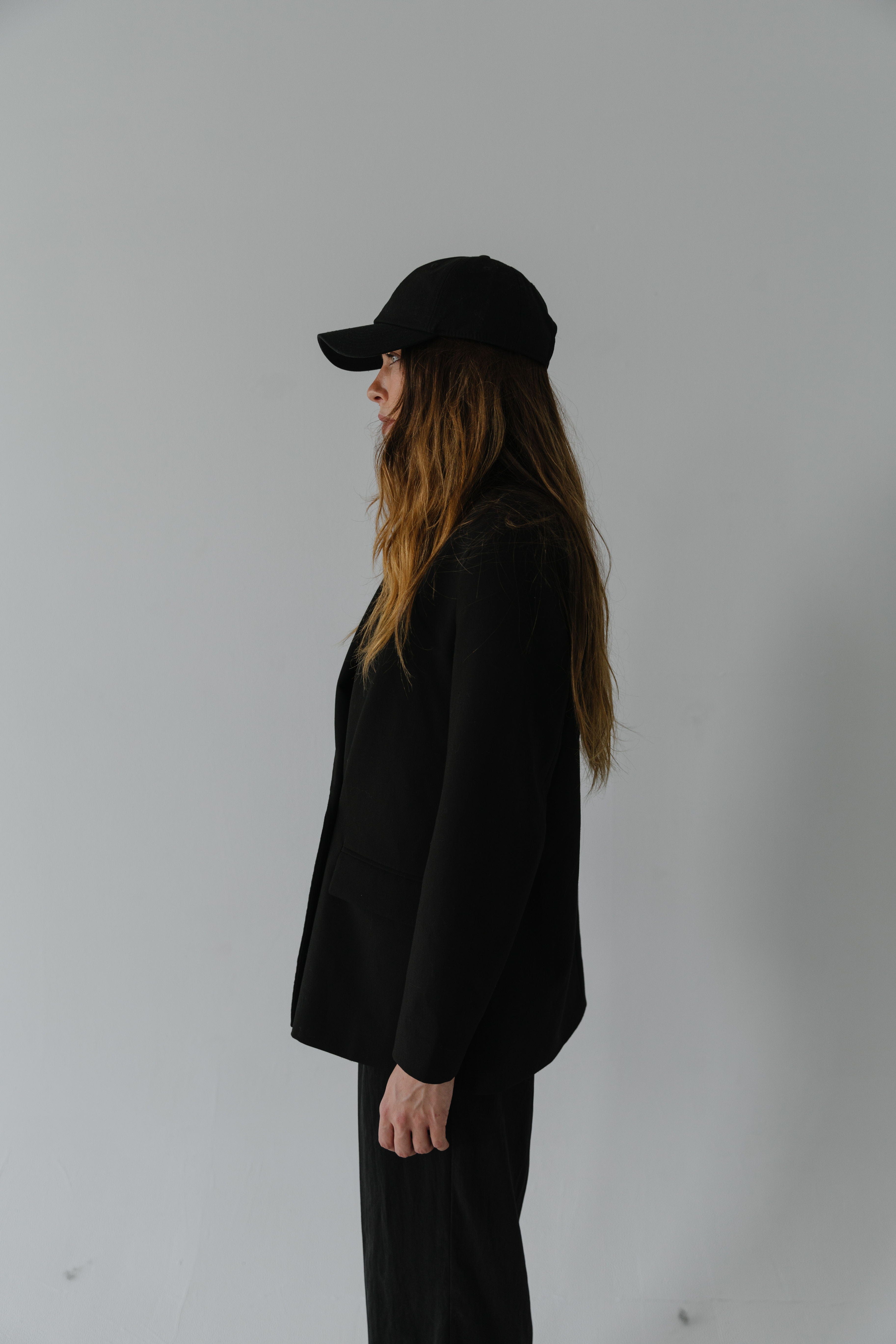 Woman wearing a black ball cap, a black coat and black pants, facing sideways against a plain background #color_black