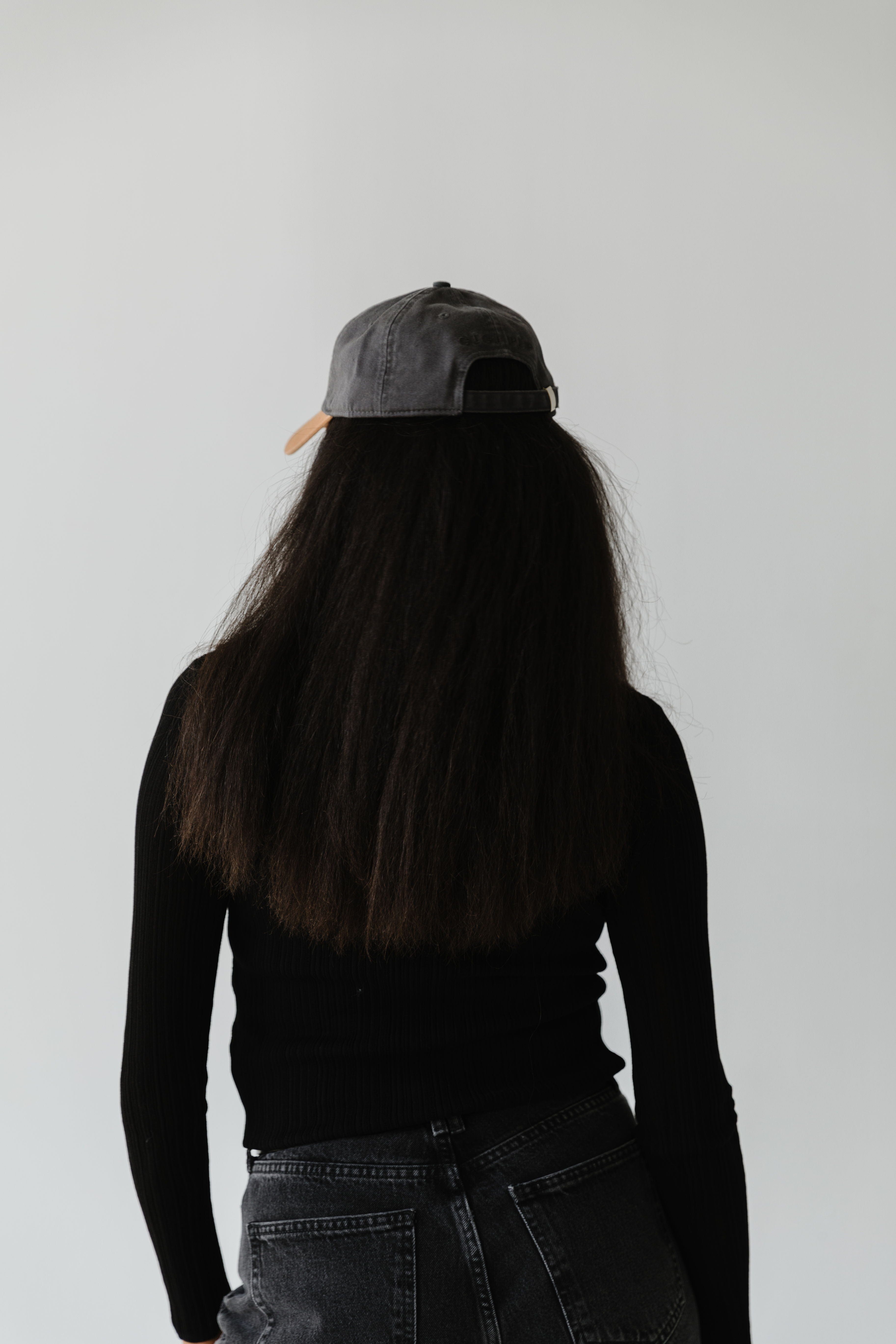 Woman with long hair wearing a dark grey and caramel colored ball cap, a black t-shirt and dark grey jeans, facing behind against a plain background #color_dark grey-caramel