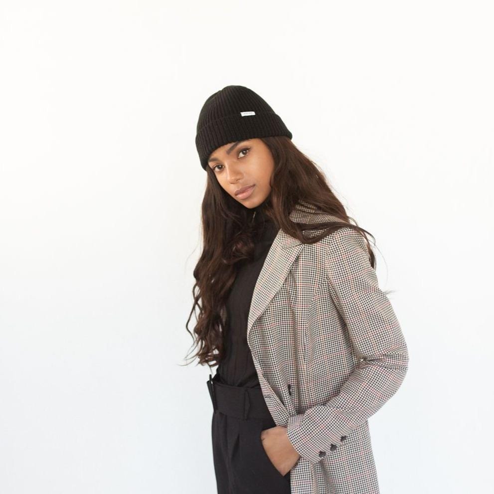 Woman wearing a black colored knit beanie with the brand label, a black top with a blazer and black pants against a plain background #color_black