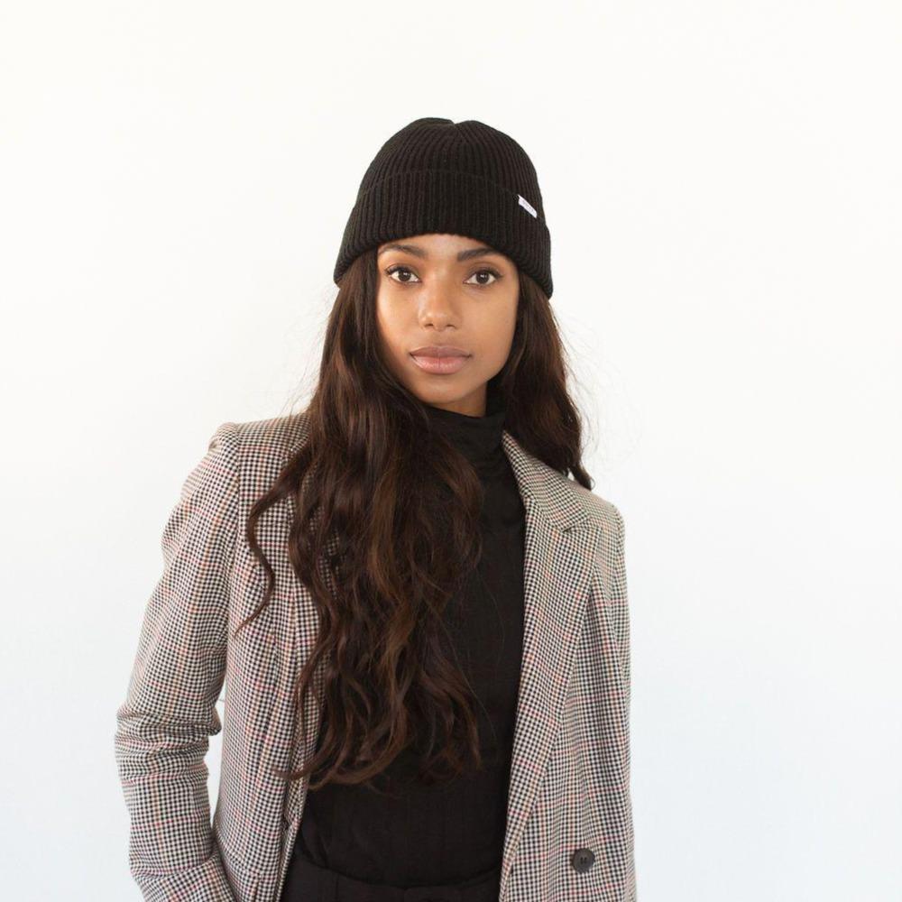 Woman wearing a black colored knit beanie with the brand label and a black top with a blazer against a plain background #color_black