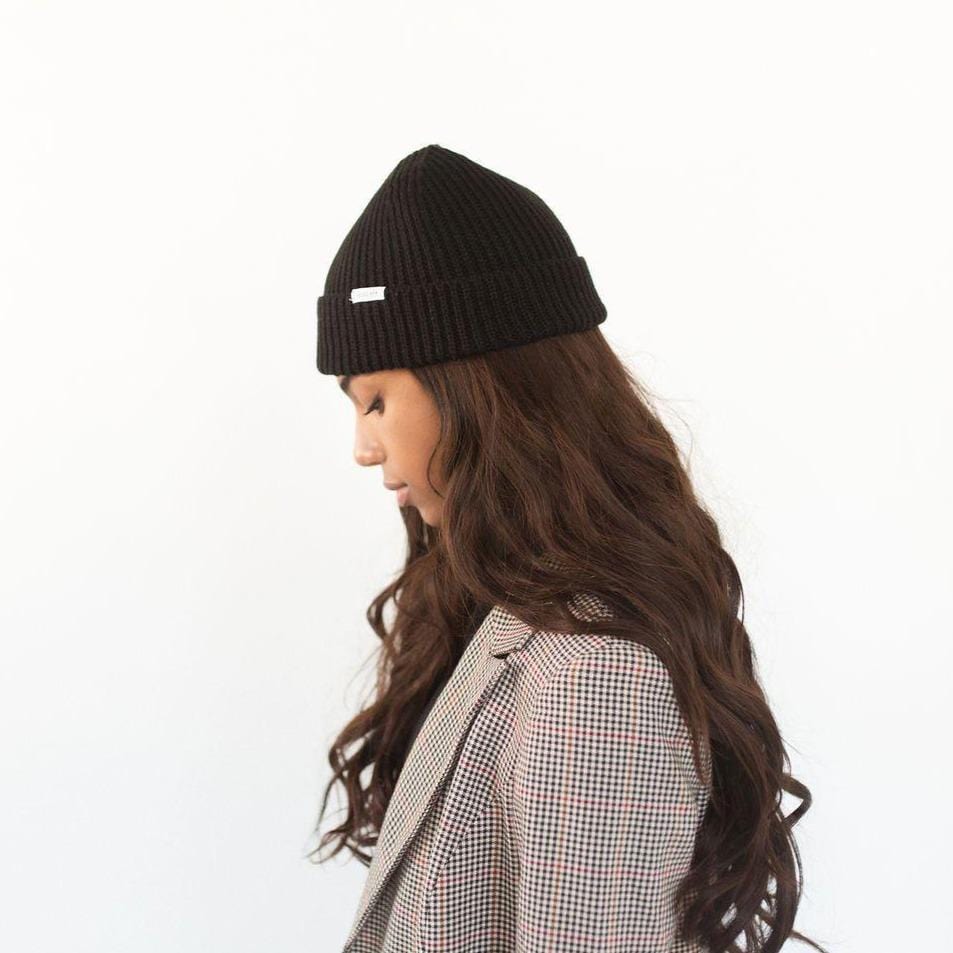 Woman wearing a black colored knit beanie with the brand label a blazer, facing sideways against a plain background #color_black