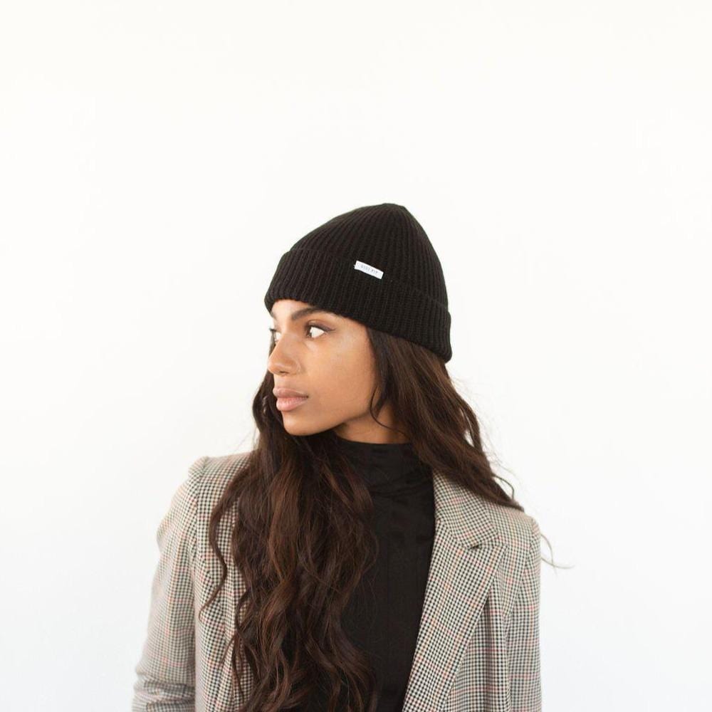 Woman wearing a black colored knit beanie with the brand label and a black top with a blazer, looking sideways against a plain background #color_black