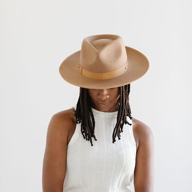 Woman wearing a brown fedora hat with a hat band and a white sleeveless top, looking down against a plain background #color_brown