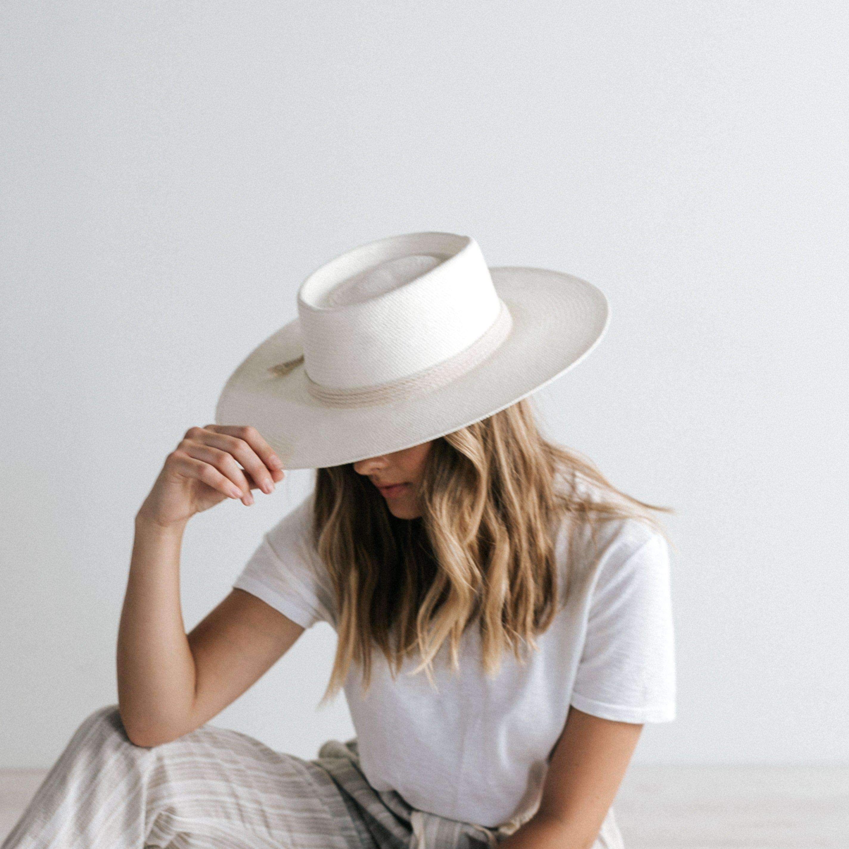 Woman wearing an ivory colored straw hat with a hat band, a white t-shirt and grey pants, posing against a plain background #color_ivory