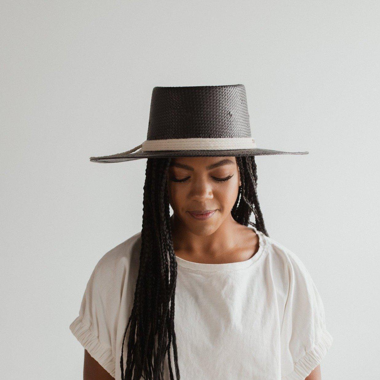 Woman wearing a black straw hat with a hat band and a white t-shirt against a plain background #color_black