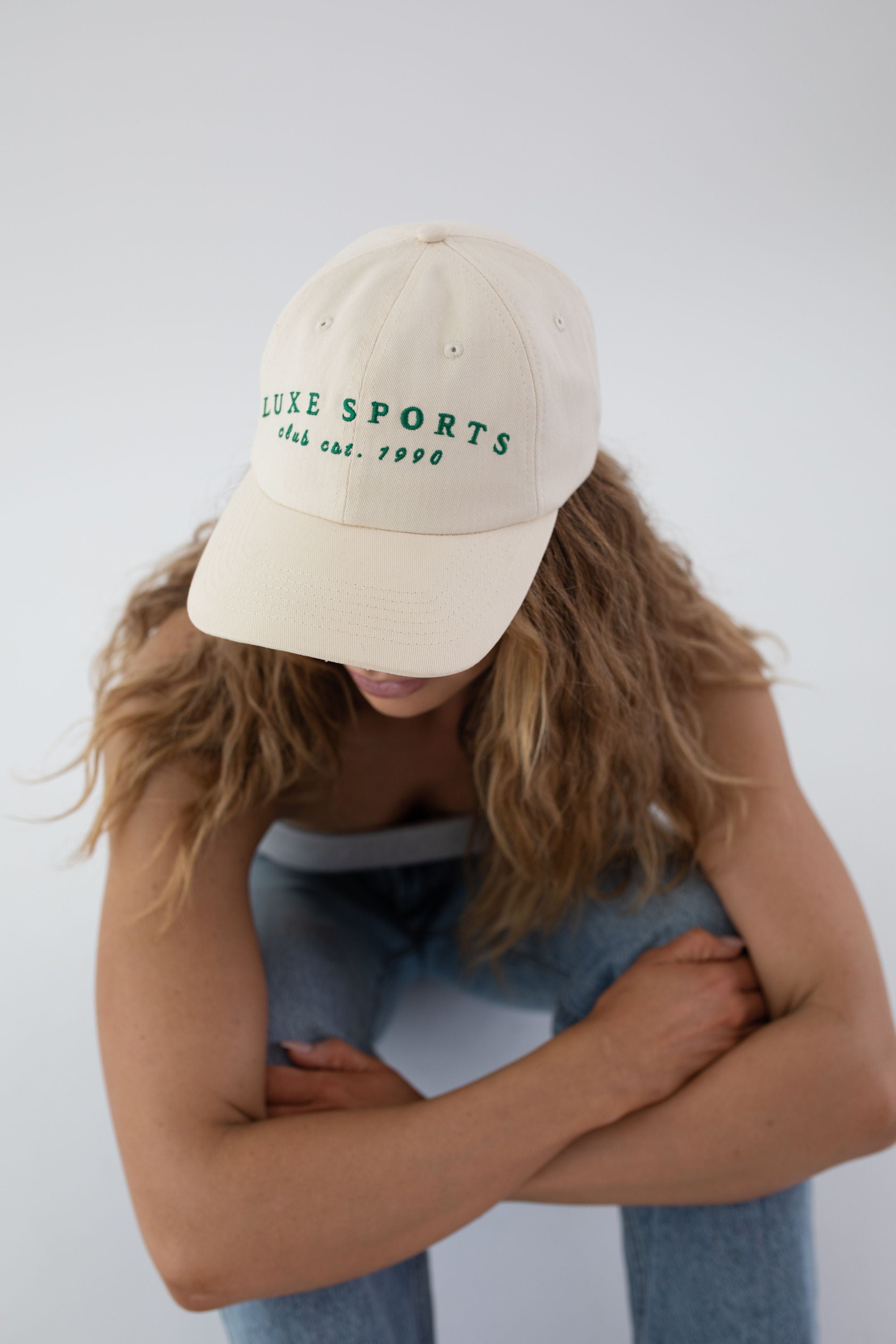 Woman wearing a cream ball cap with 'LUXE SPORTS' and 'club est. 1990' text, a white top and blue jeans, sitting against a plain background #color_cream