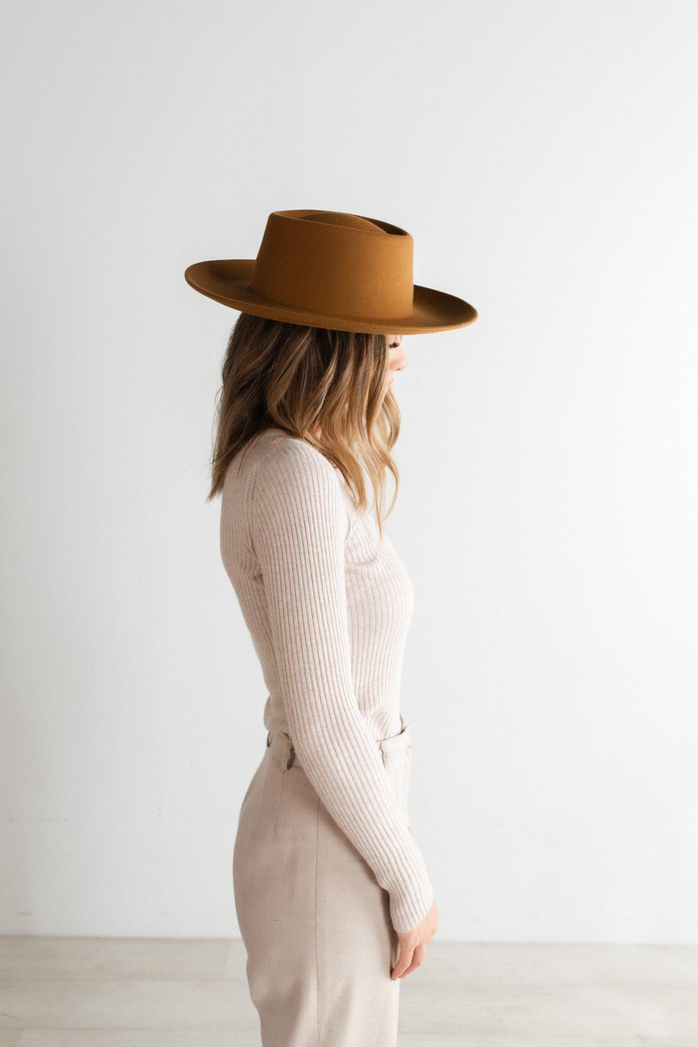 Woman wearing a cinnamon colored fedora hat, a cream top and white pants, facing sideways against a plain background #color_cinnamon