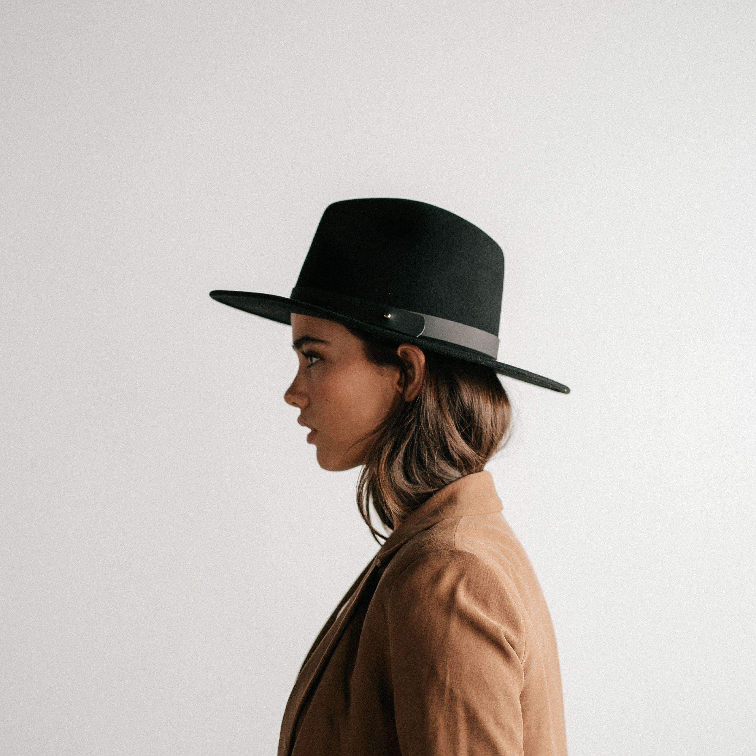 Woman wearing a black fedora hat with a hat band and a brown coat, facing sideways against a plain background #color_black