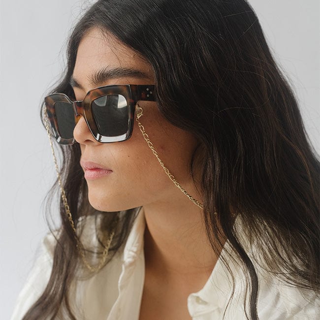 Woman wearing sunglasses with tortoiseshell frame and gold chain strap looking sideways against a plain background #color_gold