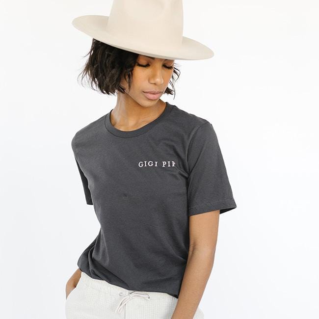Woman wearing a graphite colored t-shirt with 'GIGI PIP' text and a cream hat against a white background #color_graphite