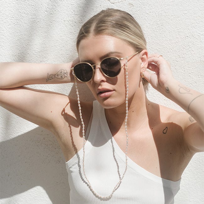 Woman wearing sunglasses with a gold pearl chain strap and a white sleeveless top against a plain background #color_gold