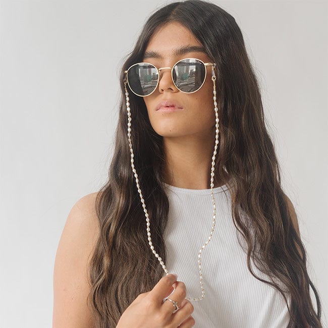 Woman wearing sunglasses with a gold pearl chain strap and a white top, looking sideways against a plain background #color_gold