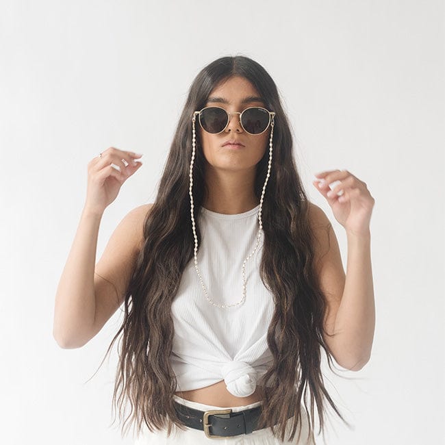 Woman wearing sunglasses with a gold pearl chain strap and a white top against a plain background #color_gold