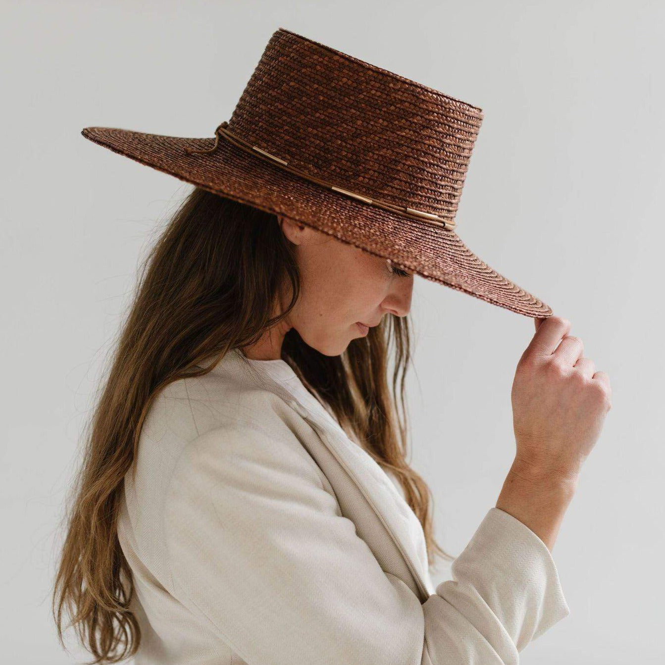 Woman wearing a brown boater-style straw hat with a flat brim and a hat band and a beige blazer, facing sideways against a plain background #color_brown