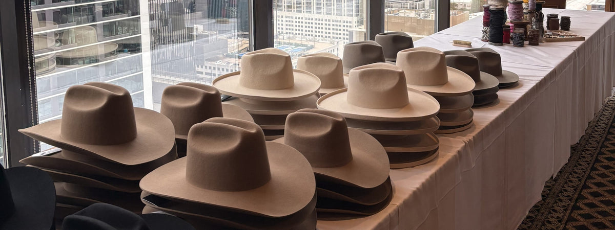 Collection of hats displayed on a table with a cityscape view through large windows at a custom hat bar event.